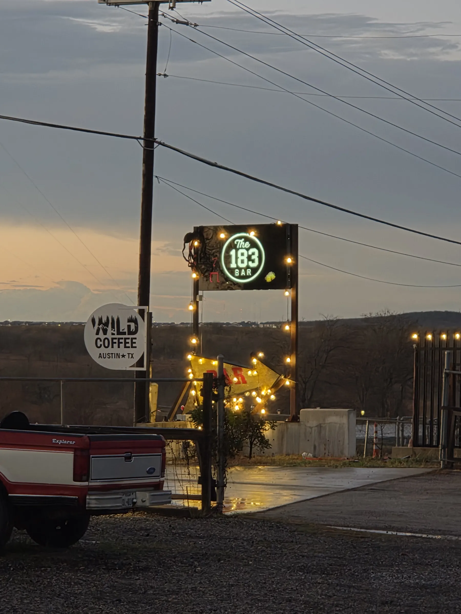 The 183 Bar sign and a bulb-lit arrow glowing at dusk beside the gravel lot off the highway