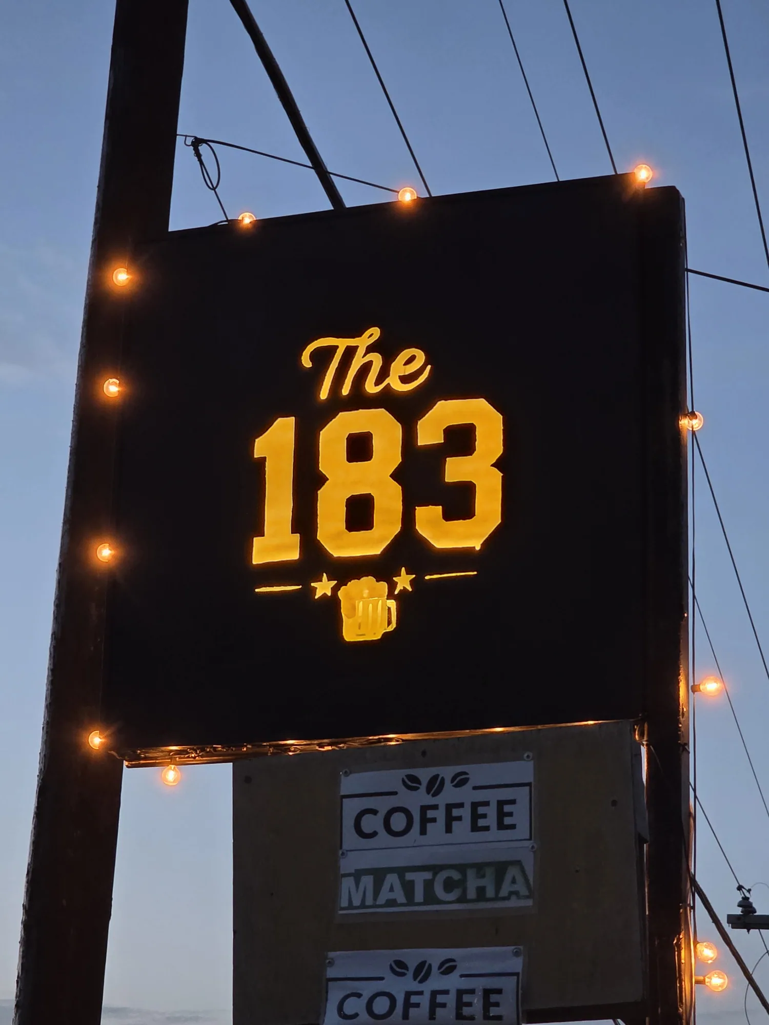 Close-up of The 183's gold lettered sign framed by glowing bulbs at dusk